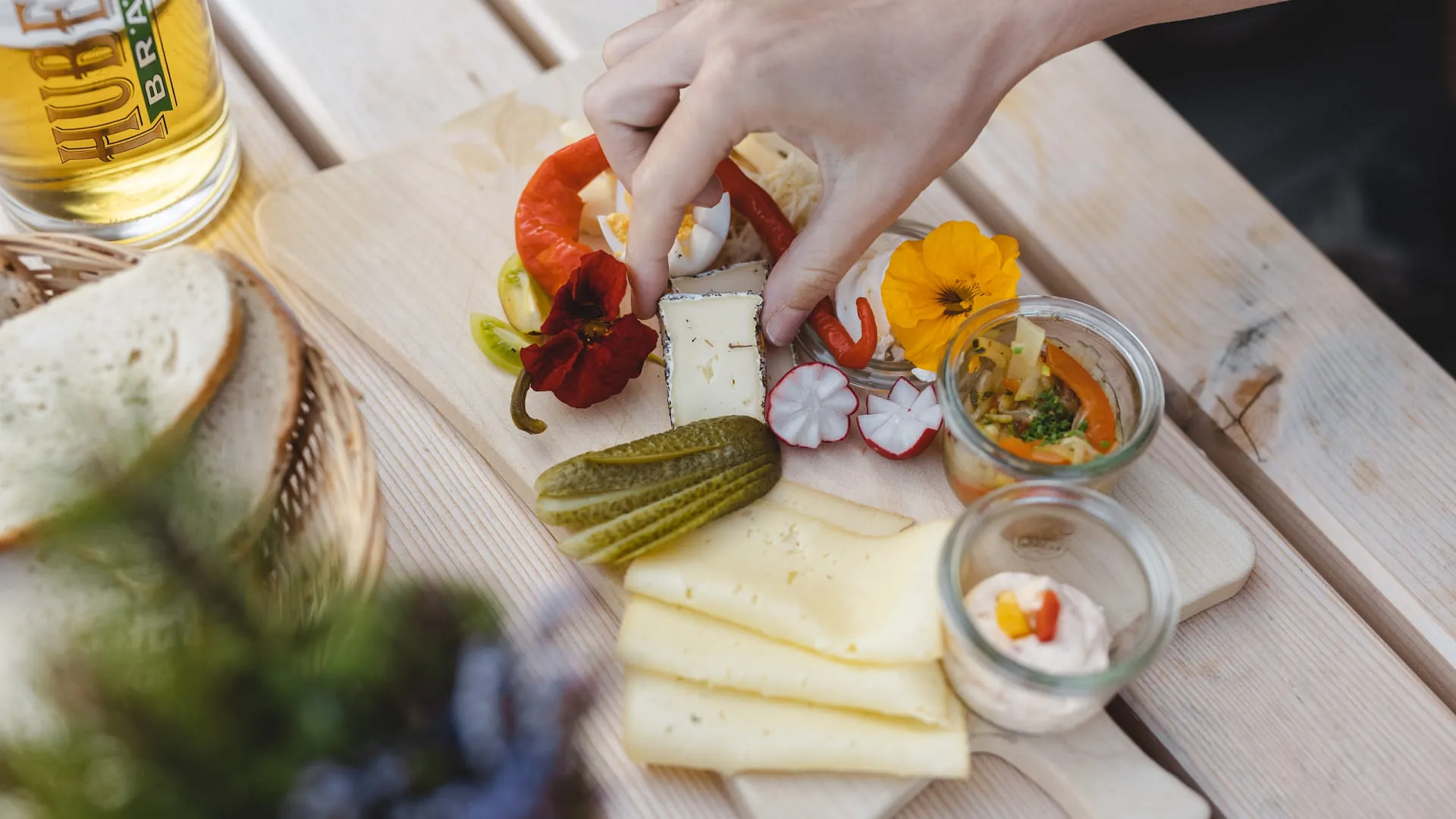 Brett mit Käse, Gemüse, Blumen und Gläsern auf Tisch.